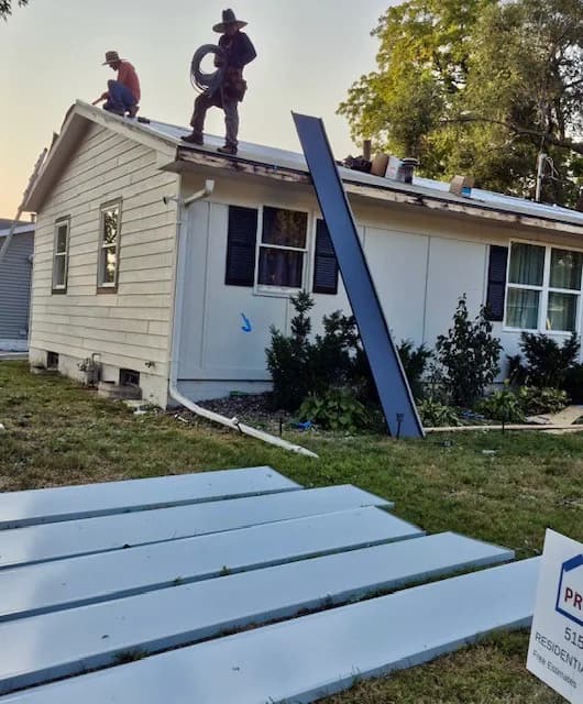 Two roofers on the ridge of a ranch house mid tear-off, with a metal panel leaning against the siding below