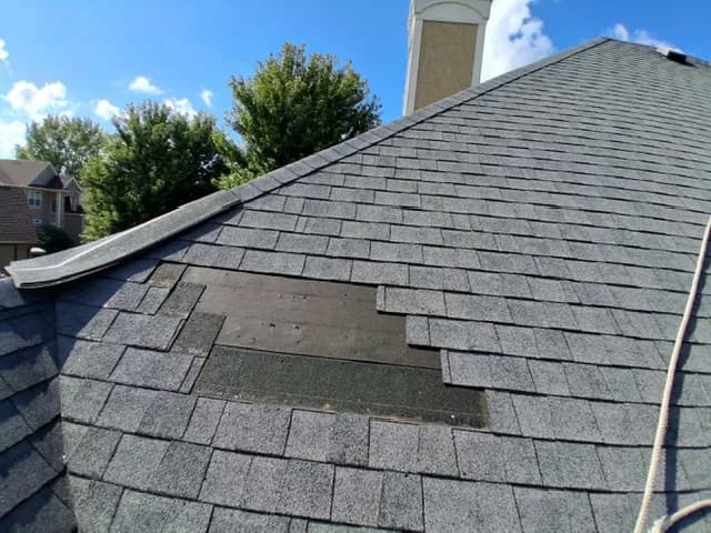 Gray shingle roof with a section of missing shingles, felt exposed underneath, chimney in the background