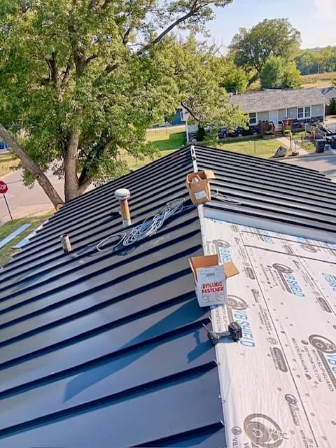 Standing seam metal panels installed halfway across a roof, synthetic underlayment visible on the unfinished half